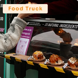 Cliente recebendo lanche em embalagem de papel em food truck, com alimentos prontos expostos sobre bancada externa.